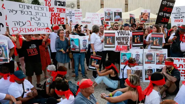 Large group of people holding signs that protest against Hamas holding hostages. Some sit on the floor with red ribbon tied around their eyes.