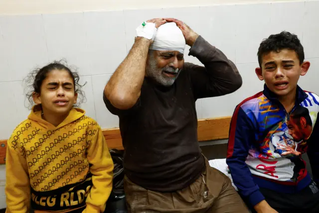 A Palestinian man wounded in an Israeli strike holds his head in anguish next to children, who also appear anguished