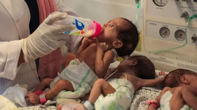 A medic out of frame supports the head of a tiny baby as it drinks milk from a bottle