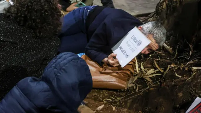 A man lies on the ground, with a sign over his face that reads: "Unicef: For every child except Israeli"