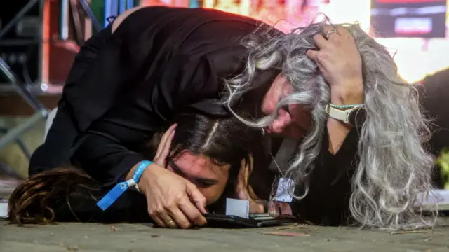 An adult with long grey hair lies prone on the ground over a teenager, who holds their hands to their ears