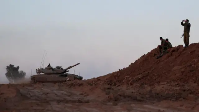 An Israeli tank manoeuvres while Israeli military serviceman watch on