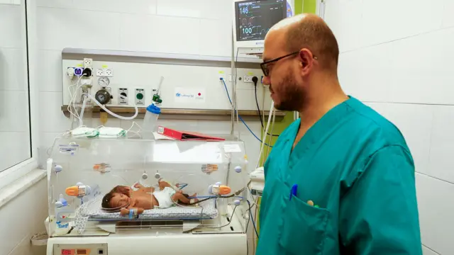 Two babies sleep in incubator as doctor looks on