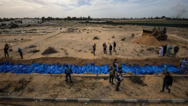 The photo is taken from above. A long trench extends the length of the frame, filled with blue body bags