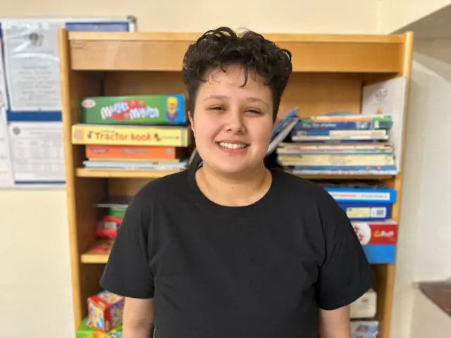 Danielle smiling for the camera, she is wearing a black T-shirt and has short, messy dark hair. Behind her is a wooden bookcase with books piled up on the shelves.