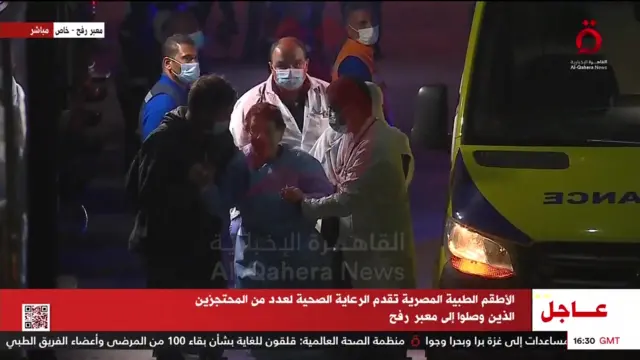 An elderly woman is shown being led into the hospital building, accompanied by men wearing medical face masks.