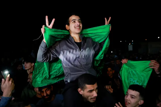 Man on another mans shoulders among crowd of people, he has a green flag draped over his back