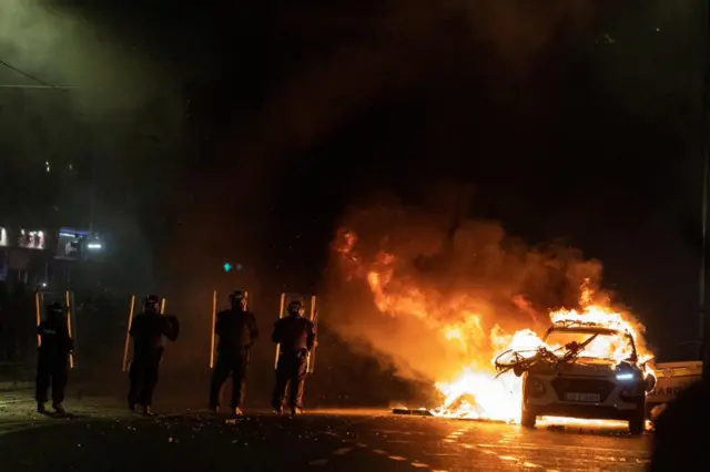 Riot police officers face down demonstrators next to a burning police car.