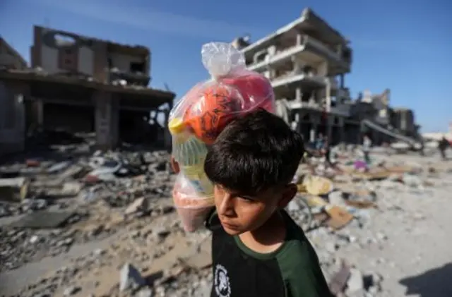 A child carrying a bag on his shoulder in front of destroyed buildings