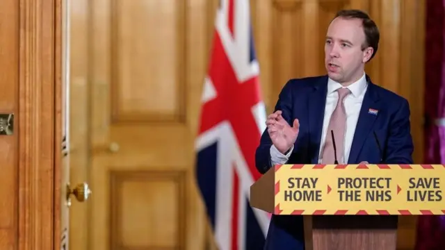 Matt Hancock speaking at a Covid conference at a plinth that says "Stay Home - Protect the NHS - Save Lives"