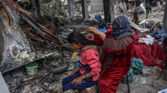 A Palestinian woman brushes a girl's hair near belongings near destoryed buildings east of Khan Younis in southern Gaza, 27 November 2023