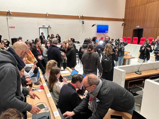 A busy court room with press standing holding camera, finding their seats etc