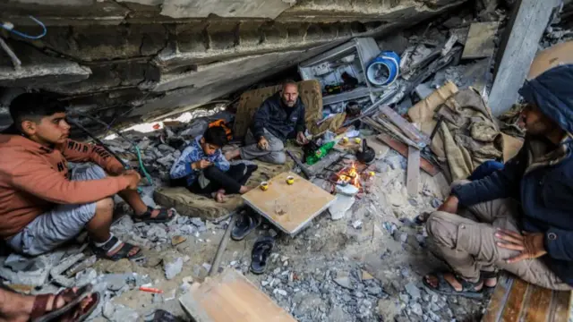 A family sitting among rubble, there is a fire with a teapot - with two men and two teenage-looking boys sitting around it.