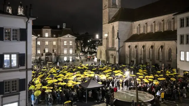 A crowd holds yellow and black umbrellas in a courtyard in Zurich