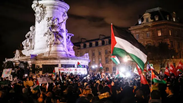 A crowd holding signs and Palestinian flags by a monument