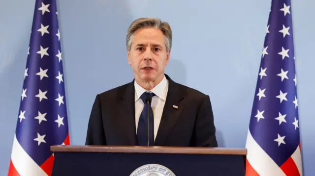 Blinken talking at a lectern, with US flags on either side