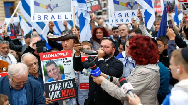 A man blows into a spiral horn, surrounded by people holding Israeli flags