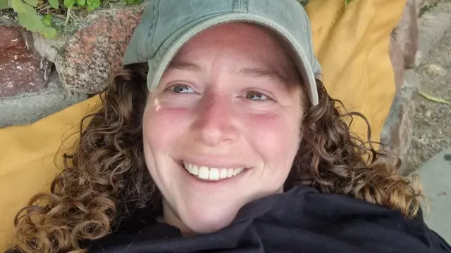 A woman with curly red hair, wearing a cap, smiles at the camera