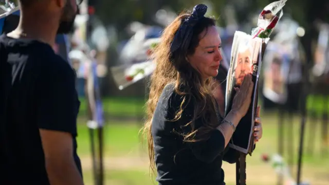 A tearful woman holds her palm against a photo of a man, one of many attached to stakes in a display