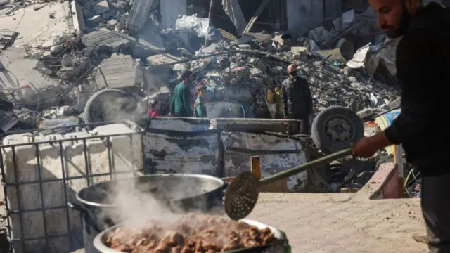 A man holds a long utensil as he watches meat cook in a large pot. In the background, people walk through rubble