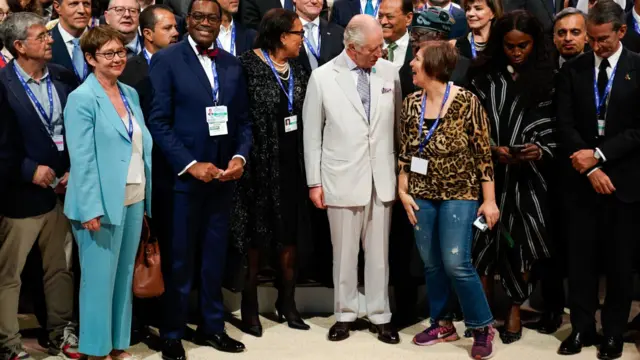 King Charles wears a cream suit as he attends the COP28 summit. He is surrounded by leaders and delegates and is seen laughing with a member.