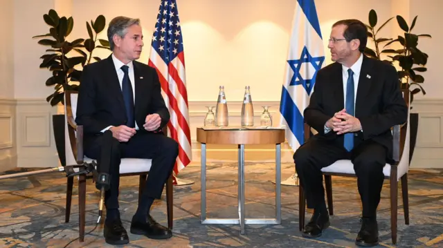 Anthony Blinken and Isaac Herzog sitting around a small round table, with the US and Israeli flags in the background