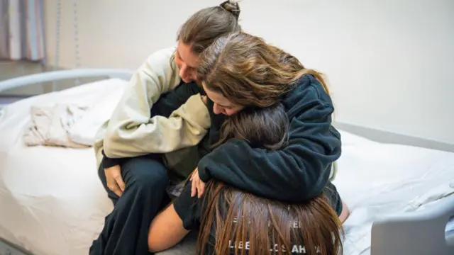 A woman sits on a bed, hugging two girls tightly