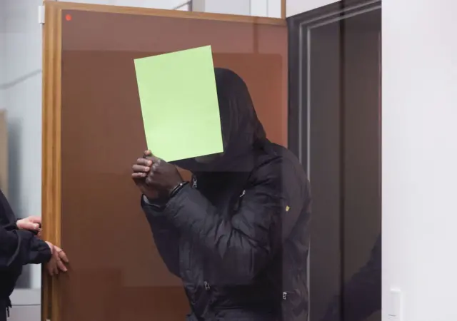 Gambian defendant Bai L, accused of of crimes against humanity, murder and attempted murder, hides behind a folder as he arrives in the courtroom on April 25, 2022 in Celle, northern Germany,