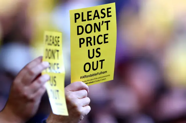 A fan holds up a card reading 'Please don't price us out [of the match]'