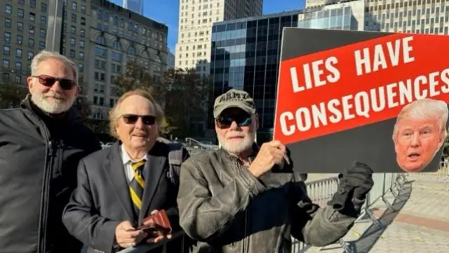 Three people stand outside Manhattan Supreme Court. One of them is holding a sign saying 'Lies have consequences' with a picture of Donald Trump's face on it