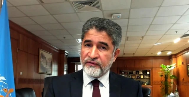 Dr Ahmed Al-Mandhari sitting in an office, a blue UN flag visible in shot. He has dark hair, a dark tie, and a beard.