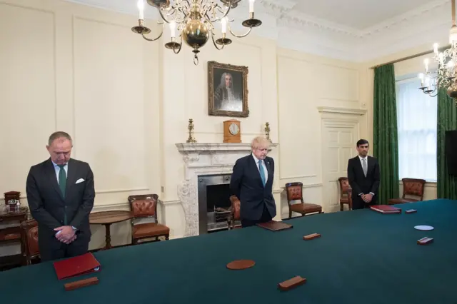 Sedwill, Johnson and Sunak stand, socially distances, with their heads bowed