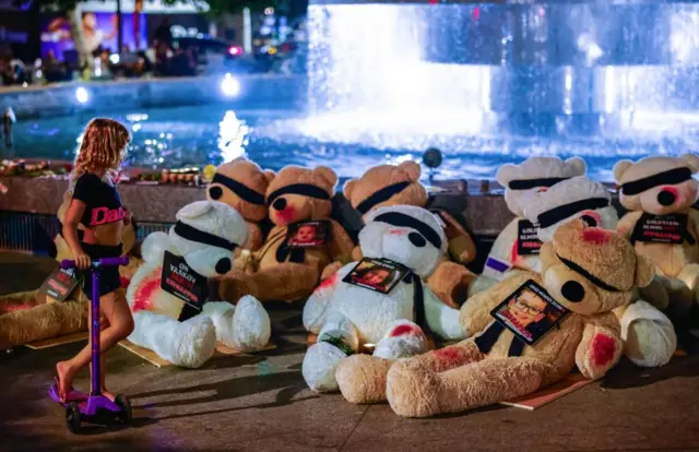 A girl stands beside teddy bears carrying the names and pictures of Israeli minors held hostage by Hamas in Gaza, displayed at Dizengoff Square in Tel Aviv