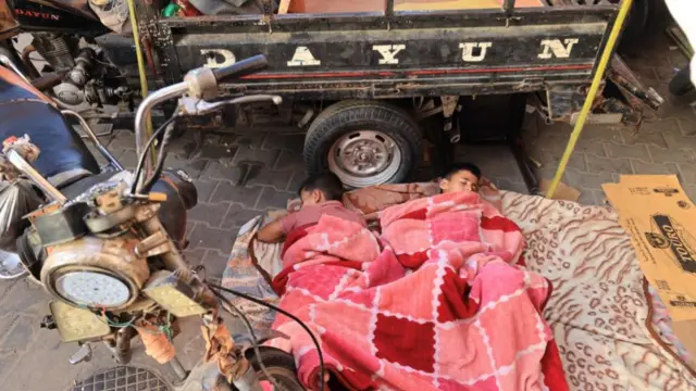 Two boys sleeping under a blanket next to a trailer and a motorbike