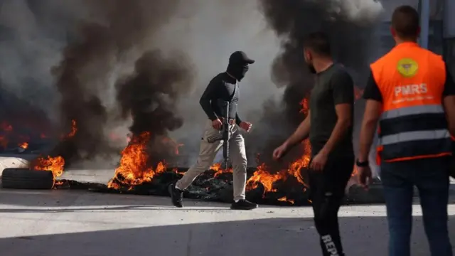 A masked militant holding a rifle walks among burning tires in Jenin. Two members look on