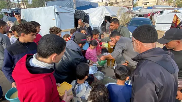 A crowd, mostly children, surround a man with a large pot of translucent yellow liquid