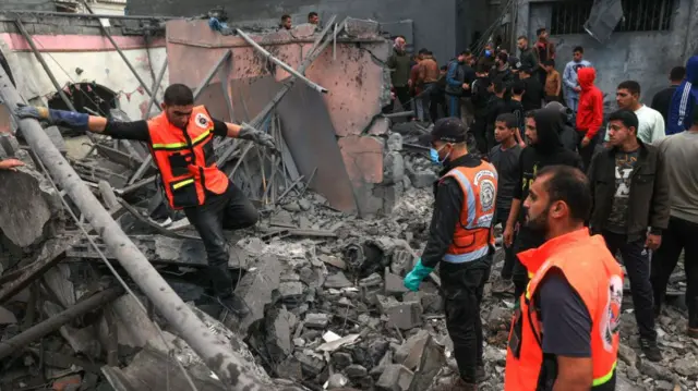 Three workers climb over rubble, as a crowd gathers behind them