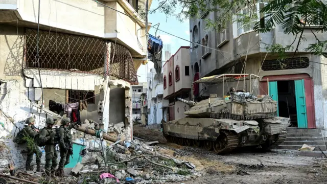 Three Israeli soldiers on foot in rubble, next to a tank operating in narrow streets in the Shajaiya district of Gaza City