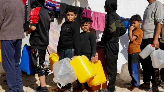 Boys, who fled their houses due to Israeli strikes, line up holding containers in a tent camp near the border with Egypt