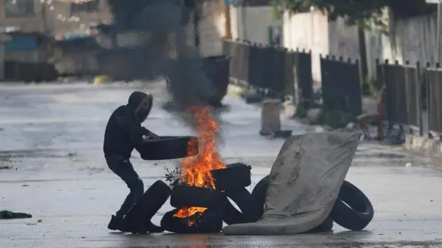 Palestinian demonstrator adds a tire to a burning makeshift road block, on the day Israeli soldiers conduct a raid in Jenin, amid the ongoing conflict between Israel and the Palestinian Islamist group Hamas, in the Israeli-occupied West Bank, December 13, 2023