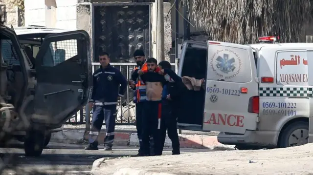 A member of an emergency medical team is checked by lifting his shirt as he stands next to an ambulance , to expose his bare chest. An Israeli armoured vehicle with its doors open is parked next to them