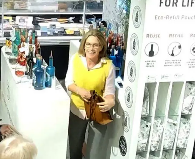 Smiling Gaynor Lord in a yellow vest over a white shirt and black trousers. She is holding a brown handbag and exiting from behind a gin counteer in Jarrolds