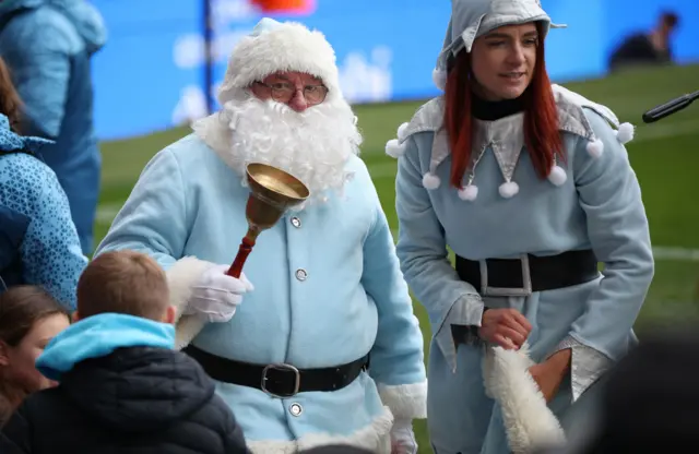 Man dressed as Santa Claus in light blue clothes ringing a bell at Manchester City, with a woman dressed as an elf in light blue clothes