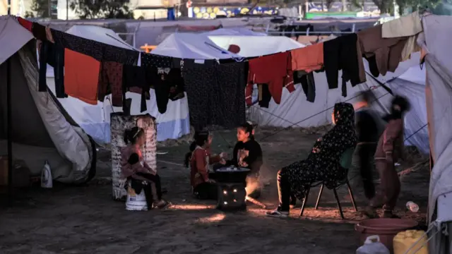 A woman sits by food cooking on a fire as children play nearby outside one of the tents housing Palestinians displaced by the conflict in Gaza