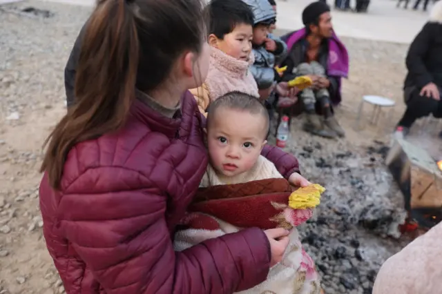 A woman wearing a puffer jacket holds an infant in freezing temperatures while sitting with others after the quake