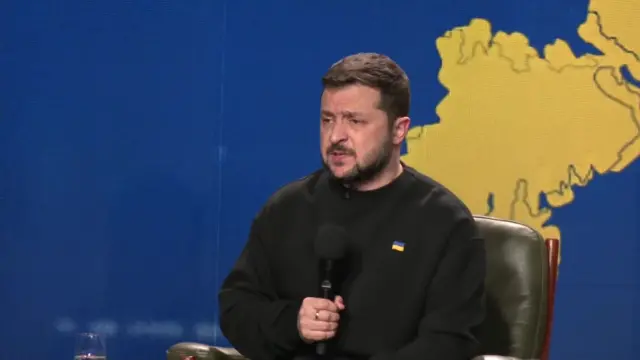 Zelensky holding a microphone and standing in front of a screen with a blue and yellow map of Ukraine