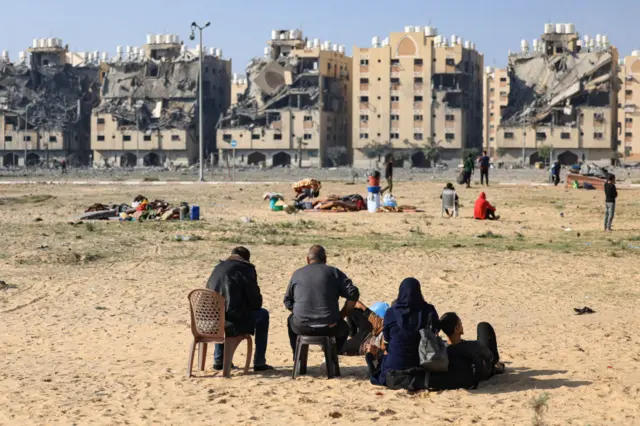 Residents sit on chairs or n the ground, looking at what's left of their homes