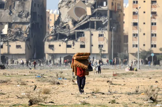 A man carries mattresses as he walks away from the ruins of Hamad Town behind him