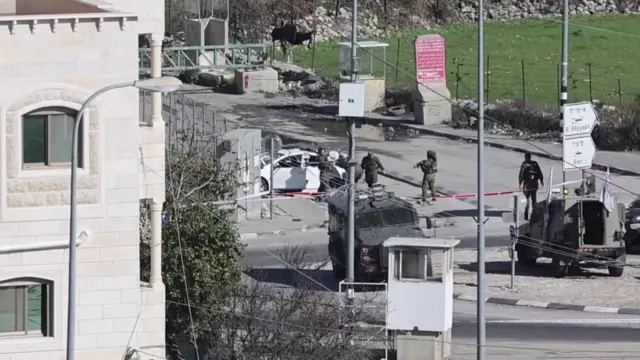 White car surrounded by soldiers, with checkpoint gate in background