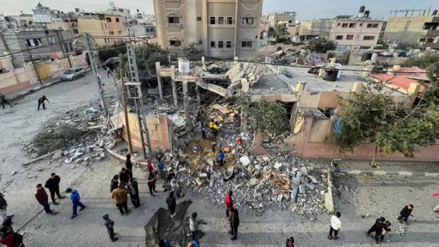 Remains of a building with rubble spreading into the street, with people standing around it